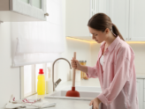 A woman stands over her kitchen sink with a plunger in hand, trying to clear a blocked drain.