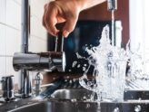 Clear water pouring from a faucet and filling a glass, with a person reaching to turn it off.