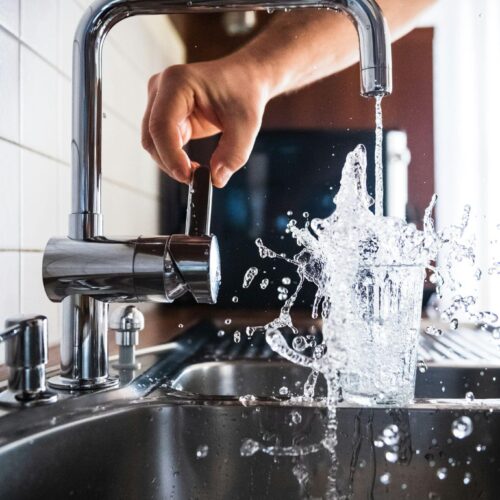 Clear water pouring from a faucet and filling a glass, with a person reaching to turn it off.