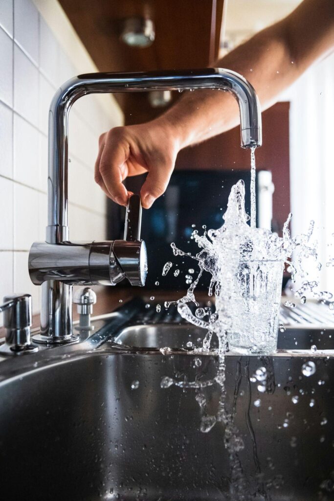 Clear water pouring from a faucet and filling a glass, with a person reaching to turn it off.